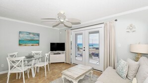 A living room with a ceiling fan, a television, a dining table with chairs, a sofa, and a view of the beach through the glass doors. - Serene Condo w/ Stunning Gulf Views on Scenic 98 in Miramar Beach! 🌊☀🏖 (Miramar Beach)