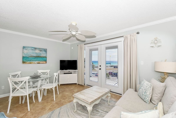 A living room with a ceiling fan, a television, a dining table with chairs, a sofa, and a view of the beach through the glass doors.