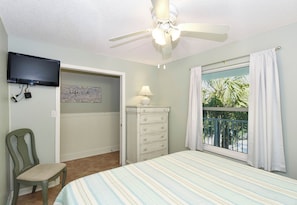 A bedroom with a bed, a ceiling fan, a television, a dresser, a chair, a window with curtains, and a view of palm trees.