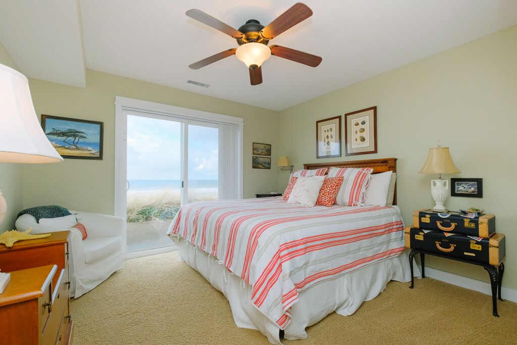 A bedroom with a bed, a ceiling fan, a nightstand, a dresser, and a window with a view of the beach.