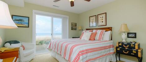 A bedroom with a bed, a ceiling fan, a nightstand, a dresser, and a window with a view of the beach.