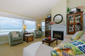 A cozy living room with a fireplace, large windows overlooking the ocean, and a collection of glassware and decorative items. - Whale watching from oceanfront home (Lincoln City)