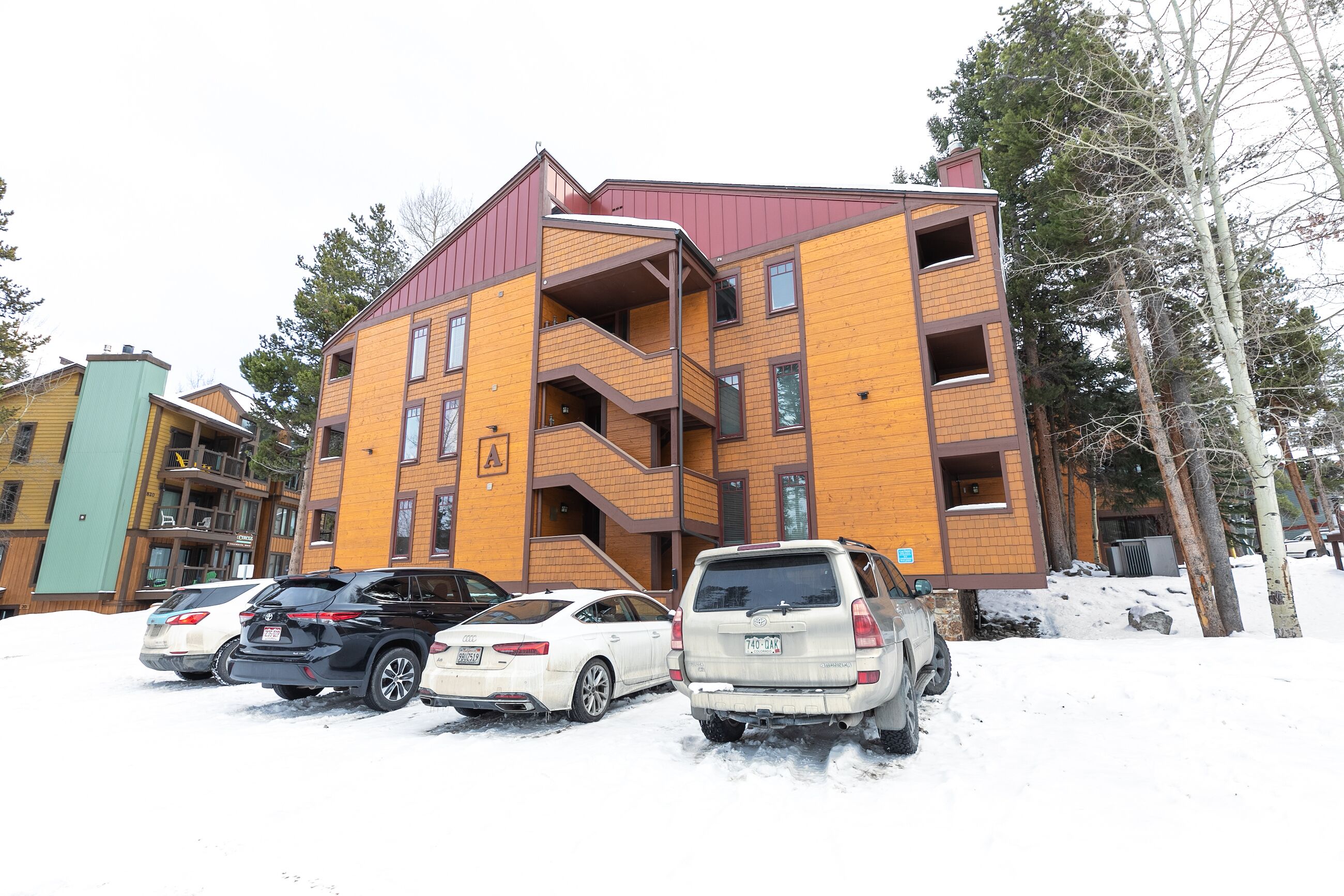Un edificio a più piani con facciata in legno e tetto rosso, dotato di un’area parcheggio con diverse automobili.