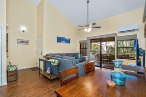 Een ruime woonkamer met een bank, eettafel en stoelen. Er is een plafondventilator, een airconditioning en uitzicht door de glazen deur.