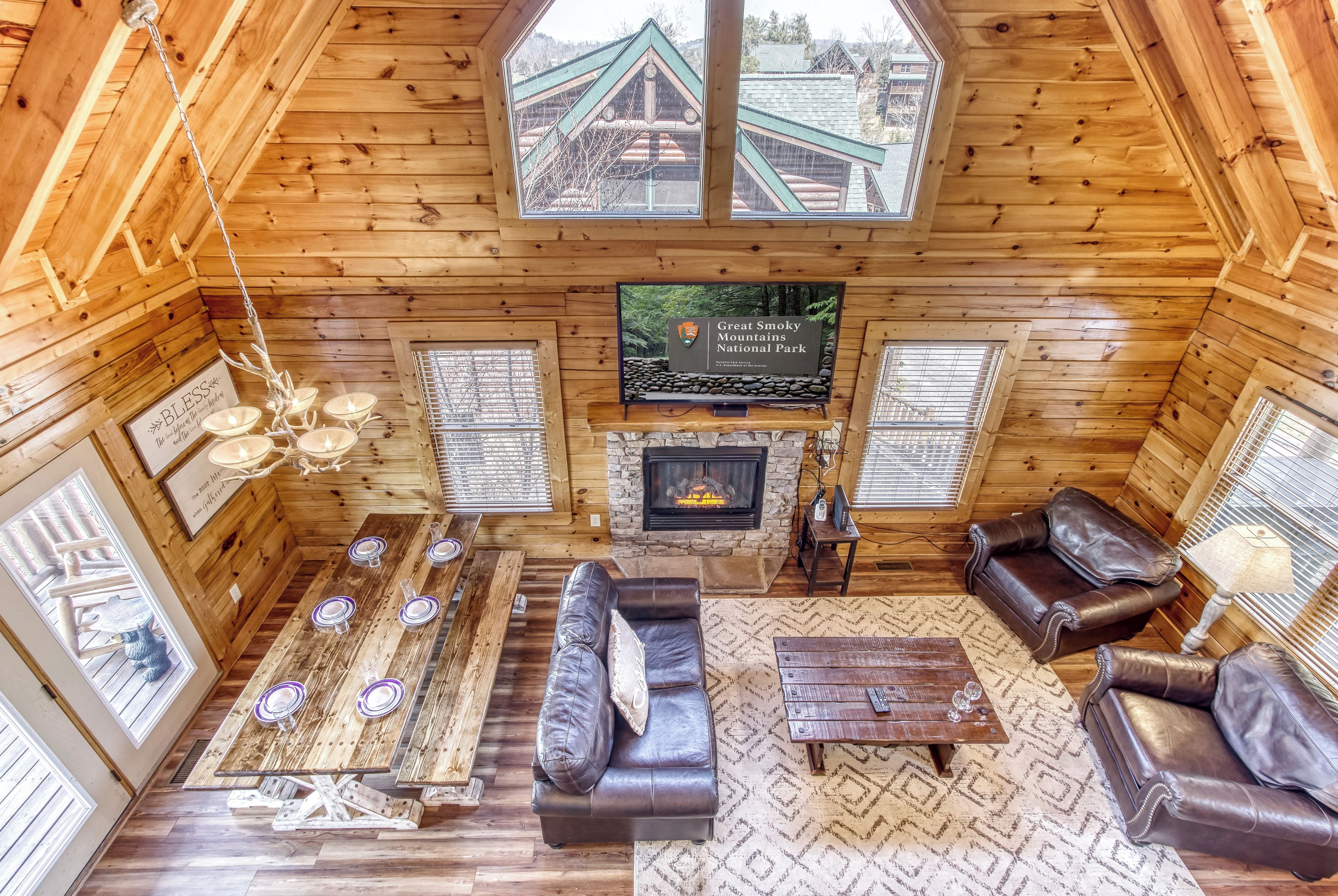 A log cabin interior with a fireplace, leather sofas, a dining table set, and a television displaying Great Smoky Mountains National Park.