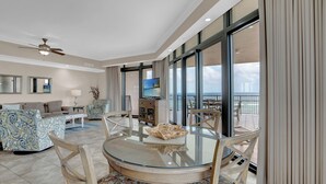A spacious living area with a glass-top dining table, comfortable seating, a television, and a balcony with a view.