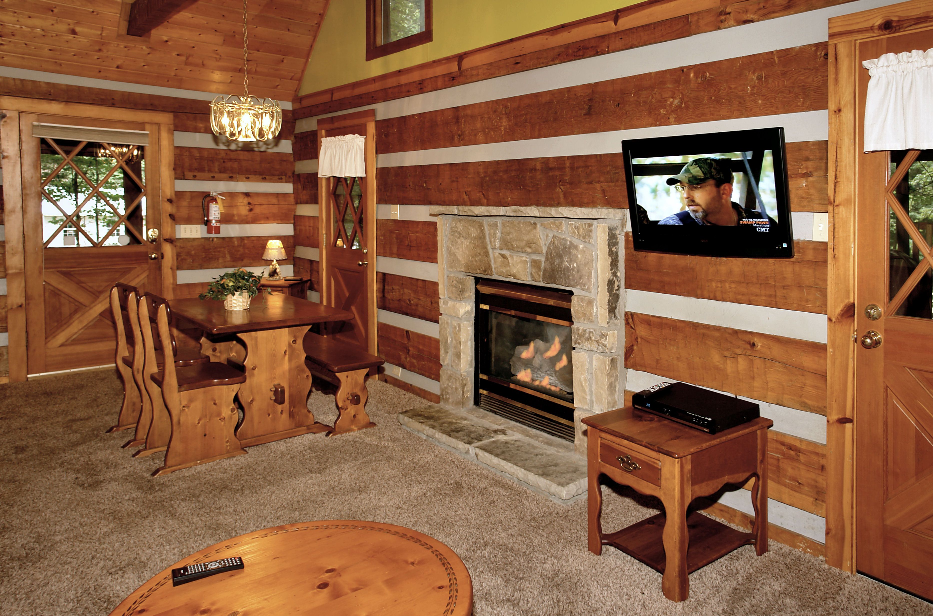 Interior de una cabaña de madera con chimenea de piedra, mesa de comedor de madera y televisor montado en la pared.
