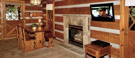 Interior de una cabaña de madera con chimenea de piedra, mesa de comedor de madera y televisor montado en la pared.