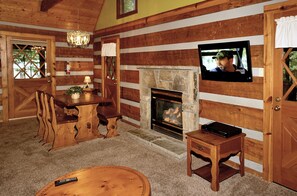 Interior de una cabaña de madera con chimenea de piedra, mesa de comedor de madera y televisor montado en la pared.