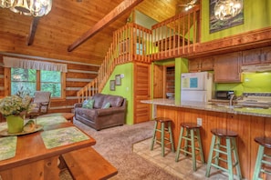 A cozy cabin interior with a wooden dining table, a sofa, a kitchen area, and a staircase leading to a loft. - Gatlinburg | Arcade | Hot Tub | Fireplace | WiFi (Gatlinburg)