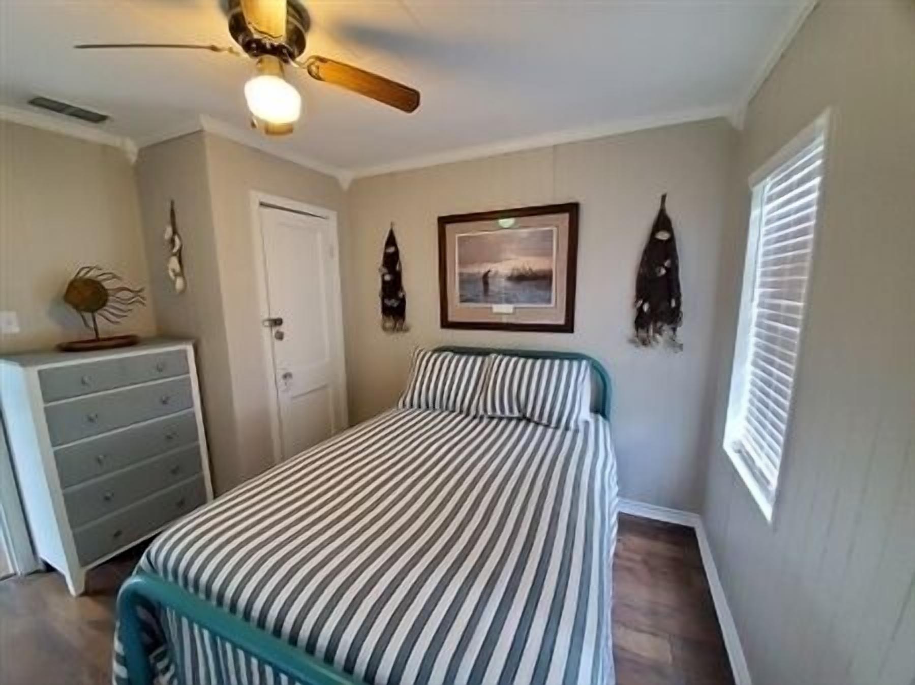 A bedroom with a bed, a dresser, a ceiling fan, a framed picture, and a window with blinds.