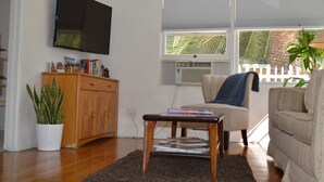 A living room with a flat-screen TV mounted on the wall, a wooden cabinet, a potted plant, a chair with a blanket, and a window with blinds.