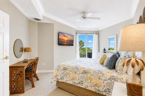 A bedroom with a bed, a desk, a chair, a ceiling fan, a window with a view, and a painting.