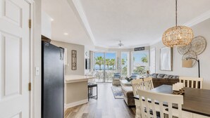 A modern kitchen with a black refrigerator, a dining area with a wooden table and chairs, and a view of the beach through large windows.