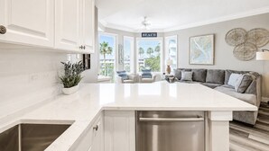 A modern kitchen with a central island, white cabinetry, and a view to a balcony with a 'BEACH' sign.