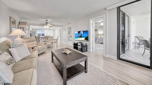 Amplia sala de estar con sofá, mesa de centro y televisor. Hay un comedor con sillas y una mesa. La habitación tiene un gran ventanal corredero que da a un área exterior con una silla.