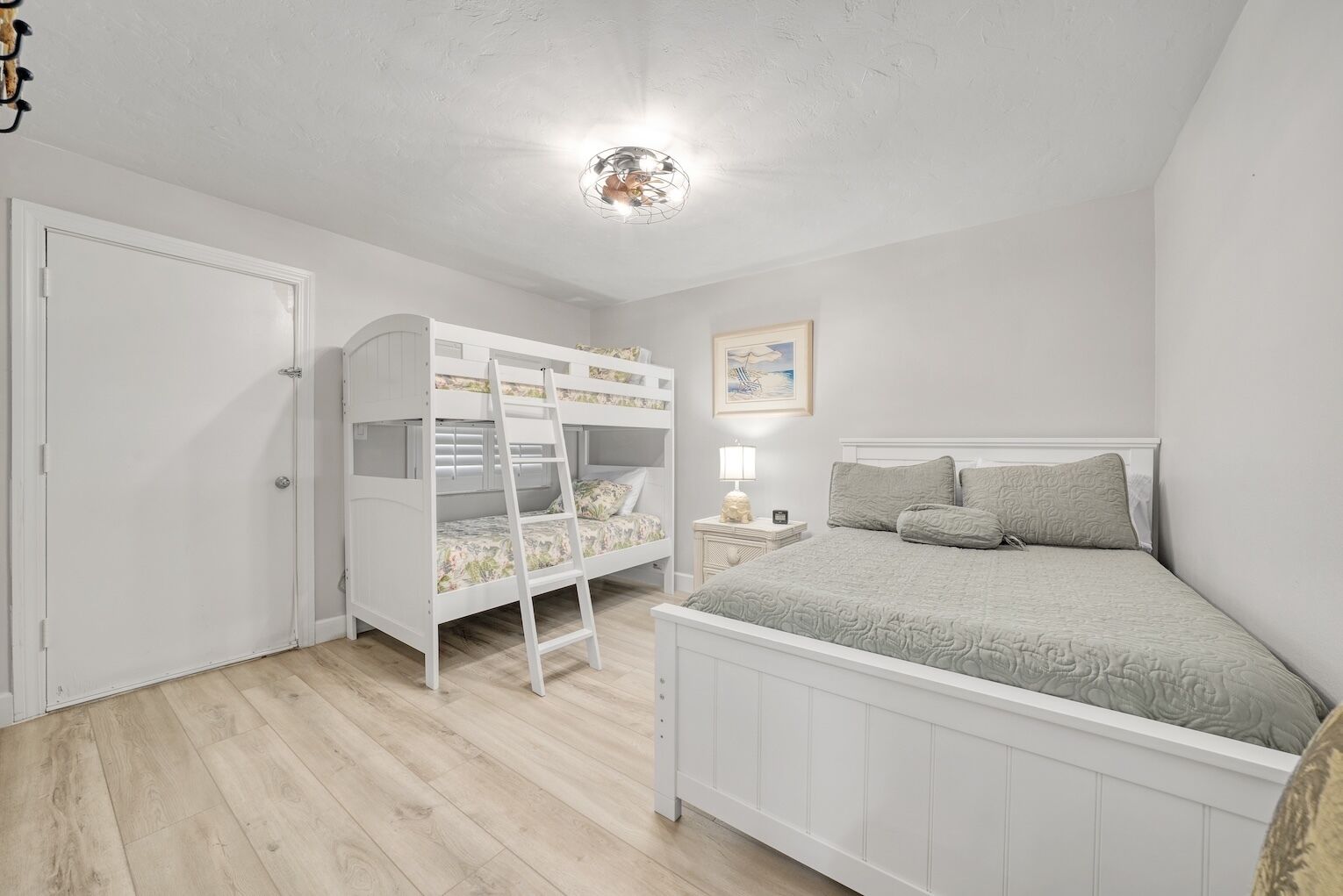 Une chambre avec un lit superposé, un lit simple, une table de chevet, une lampe et un tableau encadré au mur.