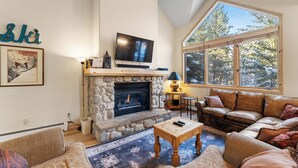 A cozy living room with a stone fireplace, a television, and a large window offering a view of trees.