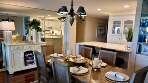 A dining area with a wooden table set for four, surrounded by chairs. There is a chandelier above the table, and a kitchen area is visible in the background. - Ocean One 413 - Oceanfront 4th Floor Condo (Hilton Head Island)