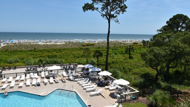 A poolside area with lounge chairs and umbrellas, overlooking a beach and ocean. - Ocean One 413 - Oceanfront 4th Floor Condo (Hilton Head Island)