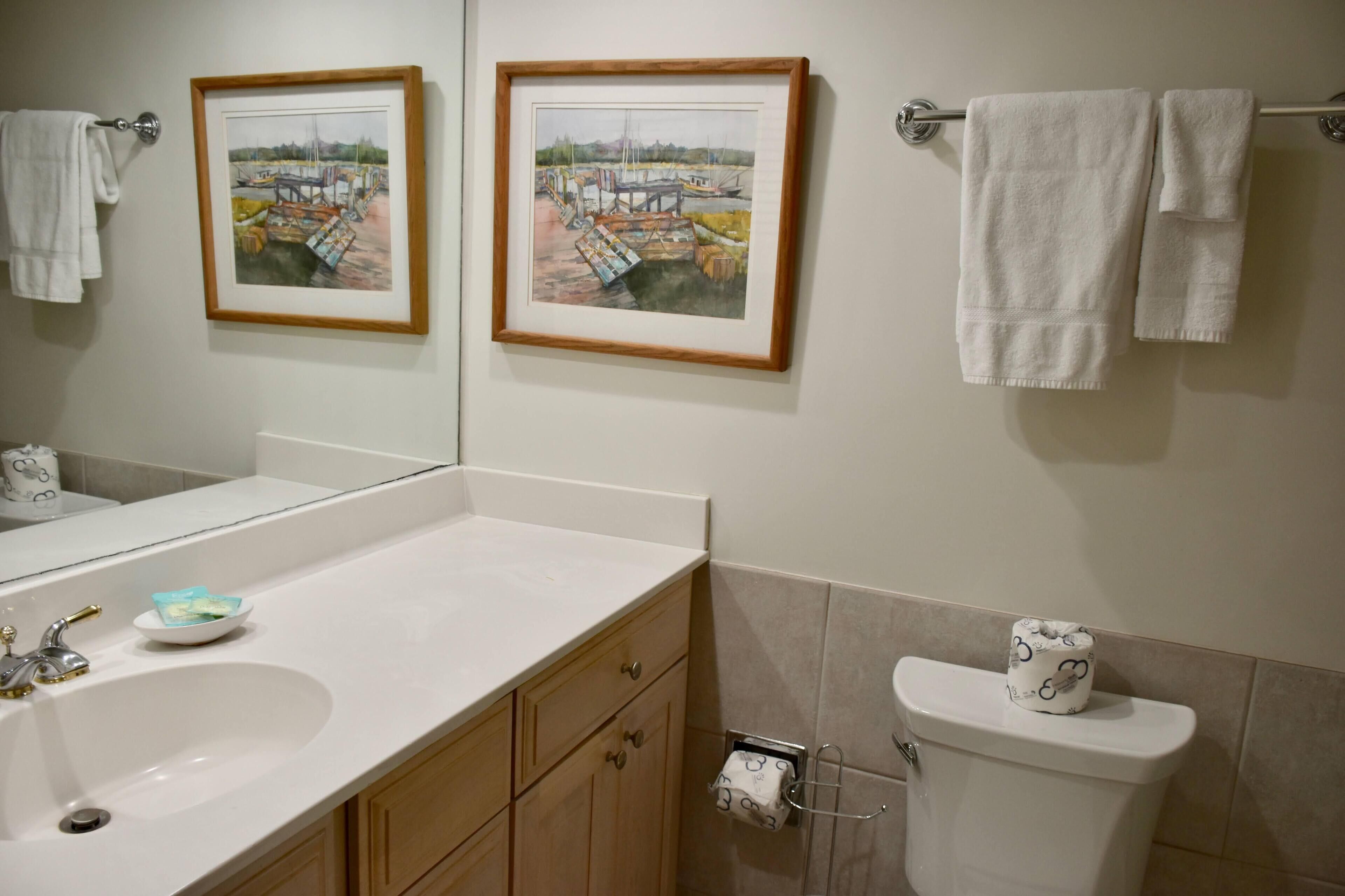 A bathroom with a white sink, wooden cabinets, a toilet with a toilet paper holder, and two framed paintings on the wall.