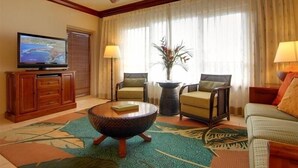 Une chambre d’hôtel équipée d’une télévision, de deux fauteuils, d’une table basse, d’un ventilateur de plafond et d’une grande fenêtre avec des rideaux.