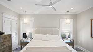 Une chambre à coucher avec un grand lit, deux tables de chevet, une commode, un ventilateur de plafond et une fenêtre avec des stores.