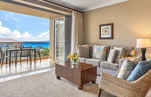 Un salon comprenant un canapé, une table basse et un fauteuil en osier, un coin repas avec une table et des chaises, et une grande porte-fenêtre coulissante donnant sur un balcon avec vue sur l’océan.