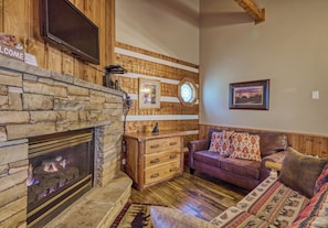 A cozy living room with a stone fireplace, wooden walls, a leather sofa, and a flat-screen TV mounted above the fireplace.