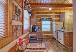 A compact kitchen with wooden walls, a refrigerator, stove, and a window with blinds.