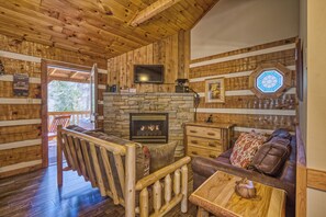 A cabin interior with a stone fireplace, wooden walls, a television mounted above it, and a cozy seating area with a leather couch and a wooden coffee table.