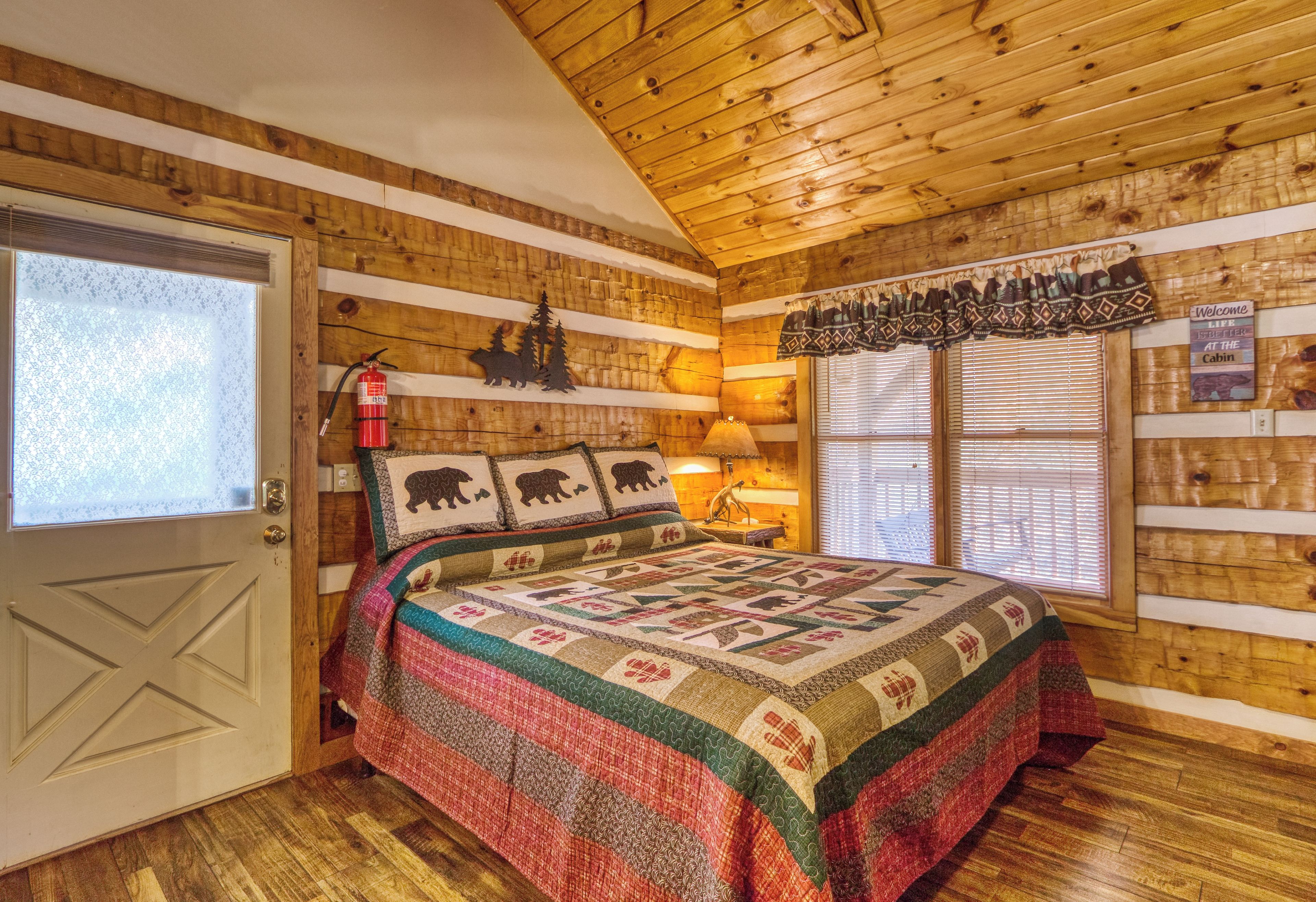 Una habitación acogedora tipo cabaña con techo de madera, una cama con colcha, un extintor y una ventana con persianas.