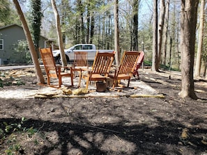 A forested area with wooden chairs and a vehicle parked near a house.