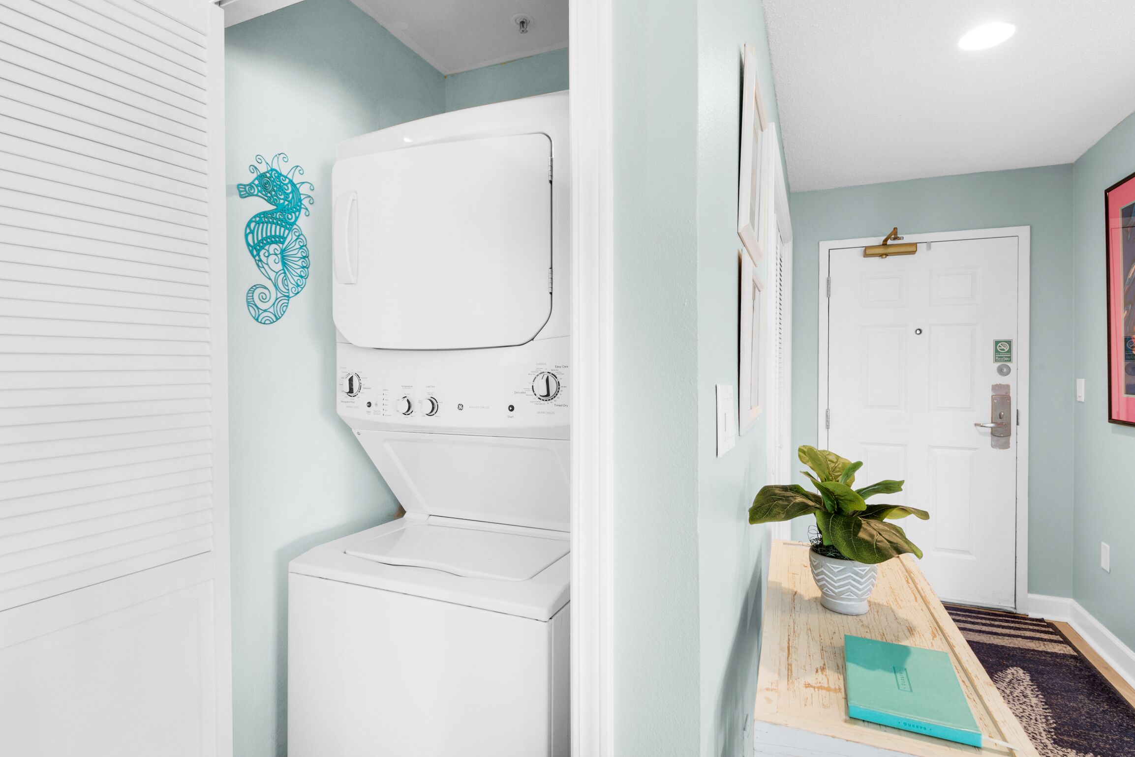 Een wasserette met een witte wasmachine, een turquoise zeepaard muursticker, en een houten bankje met een plant en een turquoise map.