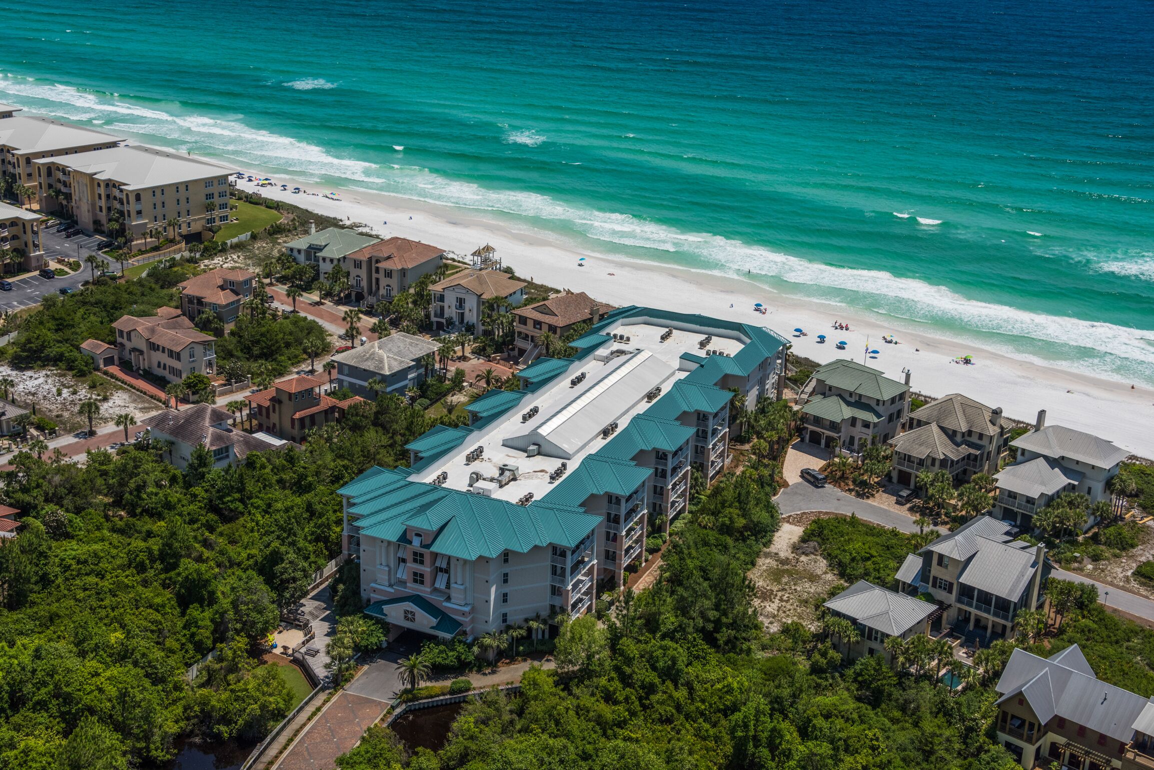 An aerial view of a coastal hotel with a large building featuring a green roof, surrounded by lush greenery and a beach.