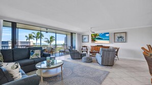 A spacious living room with a large window overlooking palm trees and a beach, furnished with a sofa, chairs, and a coffee table.