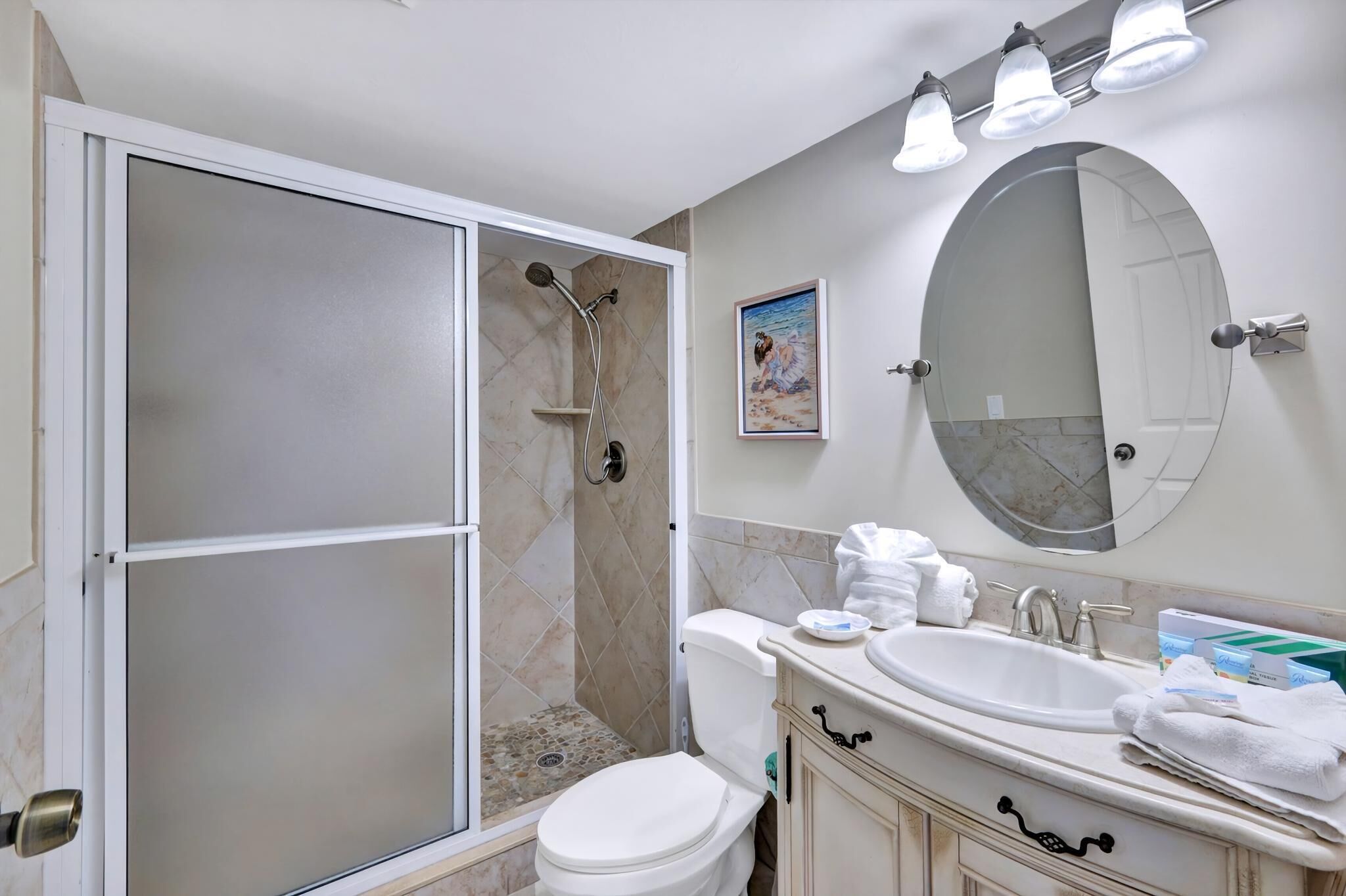 A bathroom with a glass-enclosed shower, a toilet, a sink with a mirror, and a framed picture on the wall.