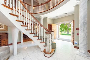 A grand staircase with white balustrades and wooden steps, leading to a room with a marble floor and a view of a green outdoor area.