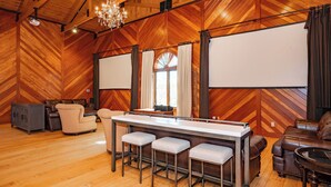 A spacious room with wooden walls and a high ceiling, featuring a bar area with stools, a sofa, and a large screen for presentations. - 20-Acre Estate w/ Pool, Games & Meeting Space (Oakland)