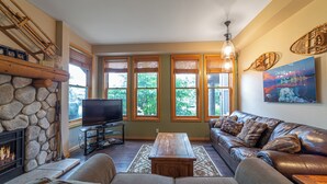 A living room with a stone fireplace, a television, a leather sofa, and large windows with blinds. - Scenic Family-Friendly Condo w/ Spa 307 (Mammoth Lakes)