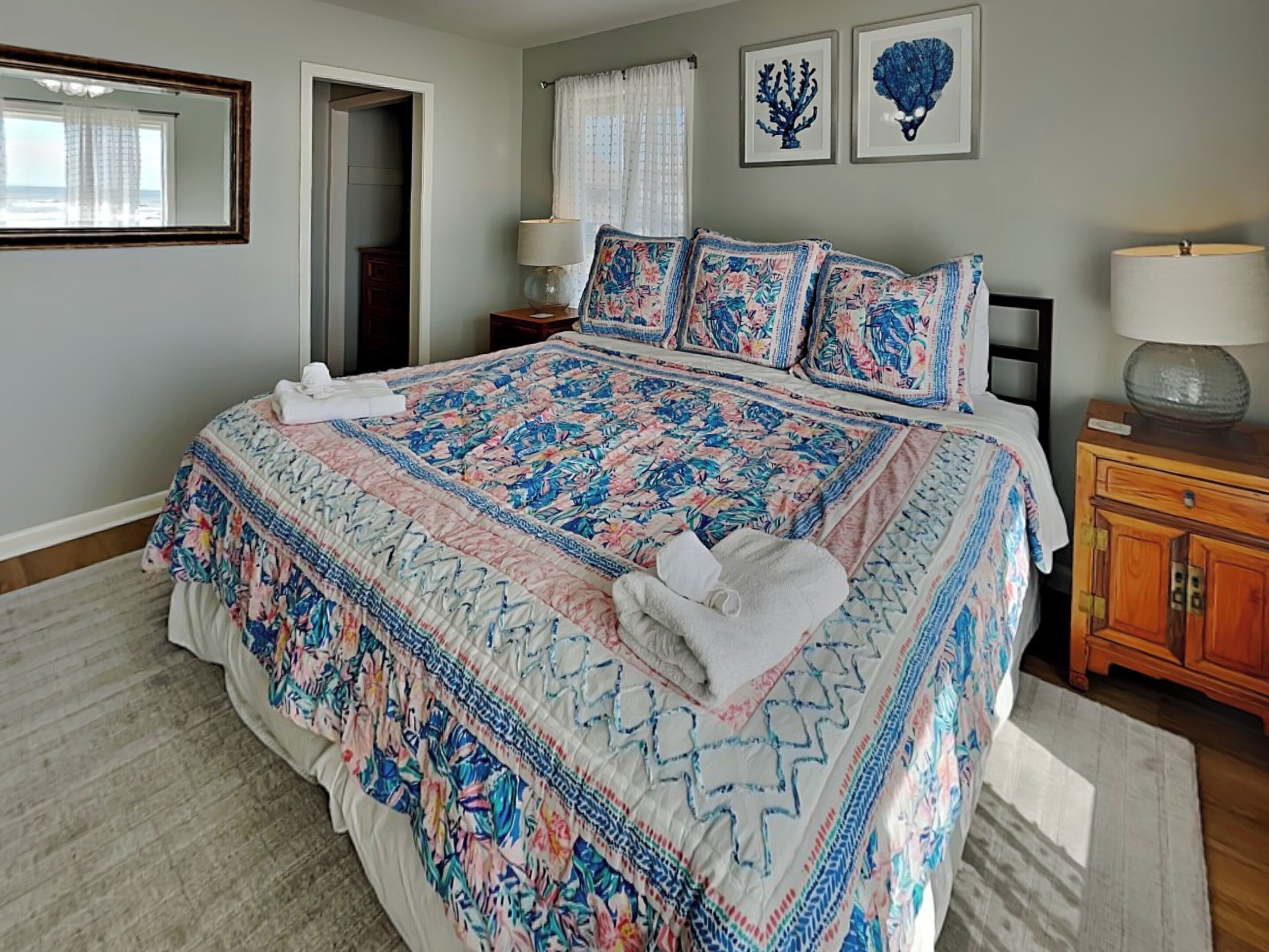 Un dormitorio con una cama grande, ropa de cama floral, una mesita de noche de madera con una lámpara y dos obras de arte colgadas en la pared.