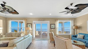 Un ampio soggiorno con un grande divano a forma di L, una poltrona blu e un tavolino da caffè. Sono presenti dei ventilatori a soffitto, un'ampia finestra con vista sull'oceano e un cartello con la scritta “BEACH”.