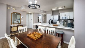 A dining area with a wooden table, white chairs, a chandelier, and a kitchen in the background. - Island Princess 509🌊Gulf Front🏖️Free Beach Setup (Fort Walton Beach)