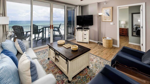 A living room with a sofa, coffee table, and TV, a balcony with chairs, and a view of the ocean. - Island Princess 509🌊Gulf Front🏖️Free Beach Setup (Fort Walton Beach)