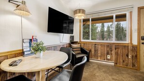 A room with a TV, a round table, and chairs, a window with a view of trees, and a wall-mounted lamp. - Economical Retreat in an Ideal Location! Ski In, Walk to Main St | PM6C (Breckenridge)