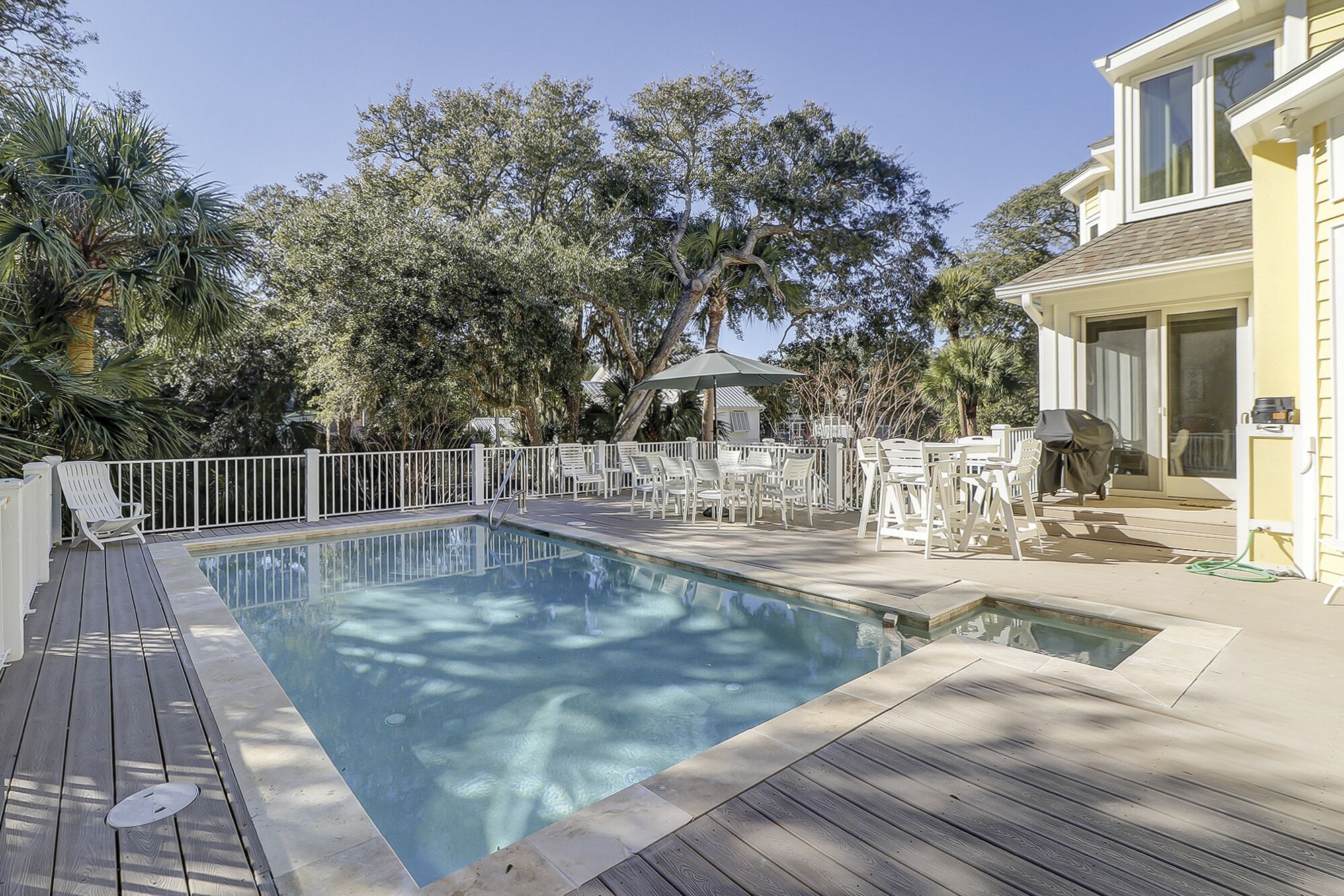 A swimming pool with a wooden deck, surrounded by white chairs and umbrellas, and trees in the background.