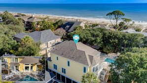 A residential area with a large house featuring a swimming pool, surrounded by trees and a beach in the background.