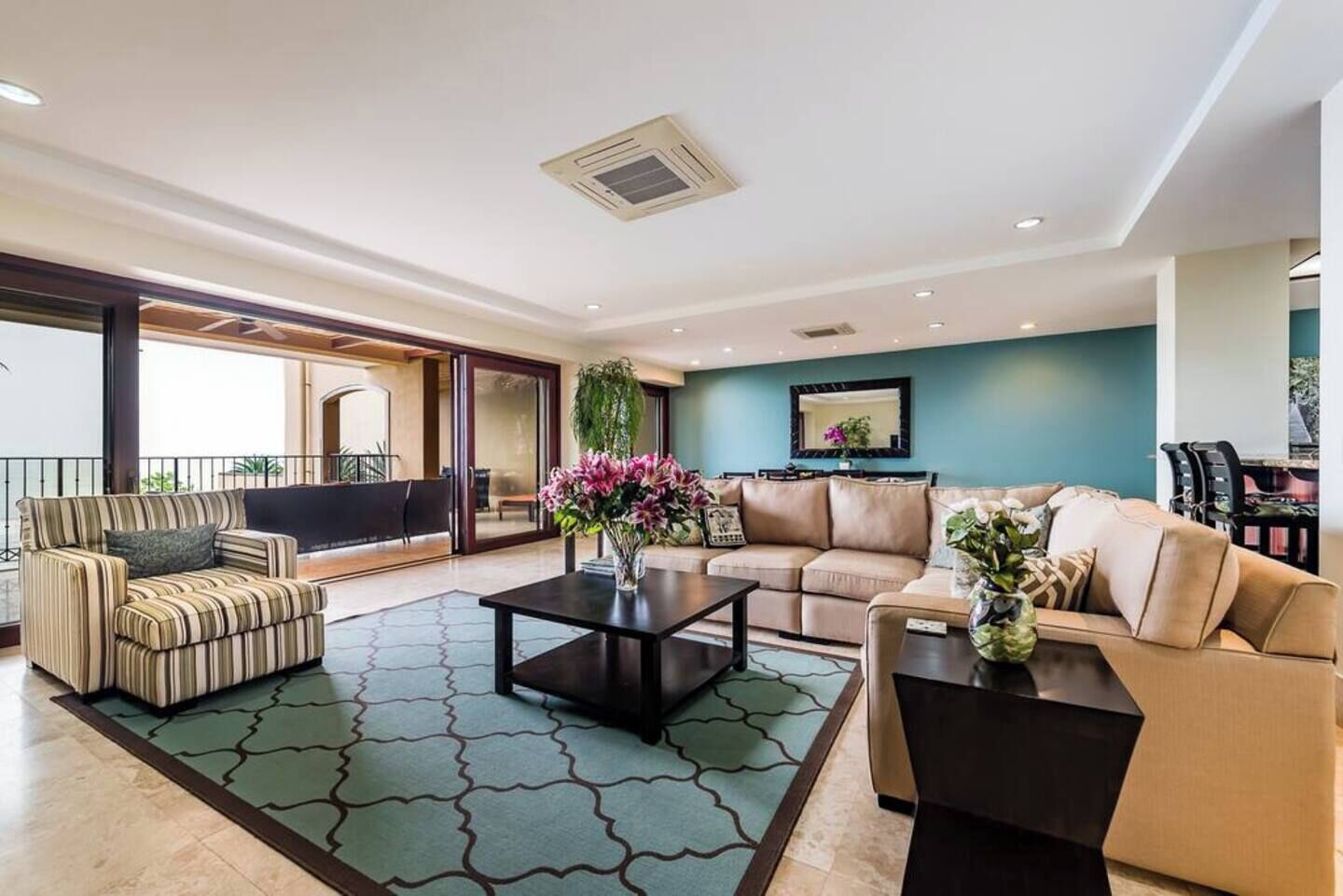 A spacious living room with a large sofa, a striped armchair, and a coffee table. There is a balcony with a railing and a view of the outdoors.