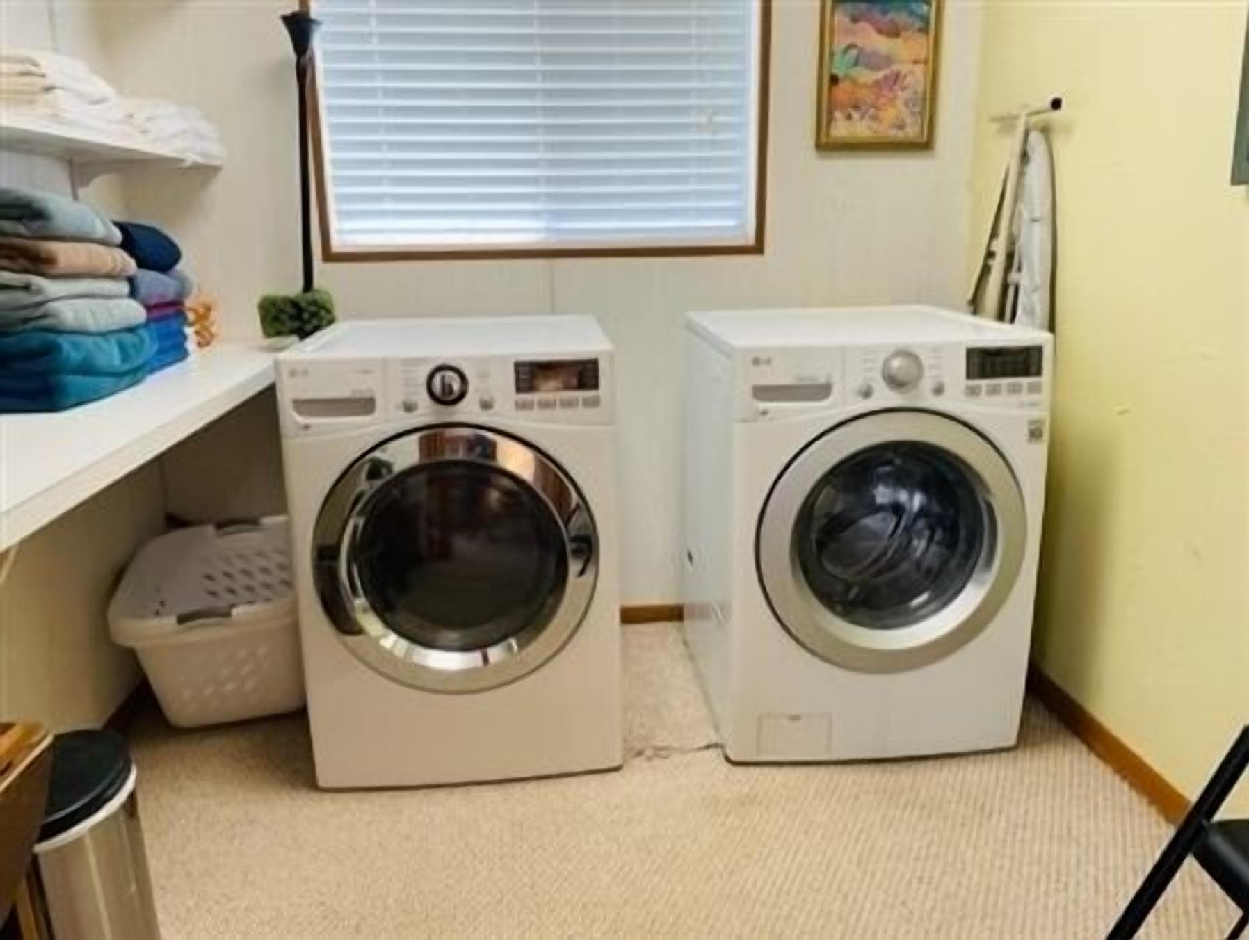 Deux machines à laver à chargement frontal se trouvent dans une buanderie équipée d’une fenêtre, d’un étendoir mural et d’une petite plante.