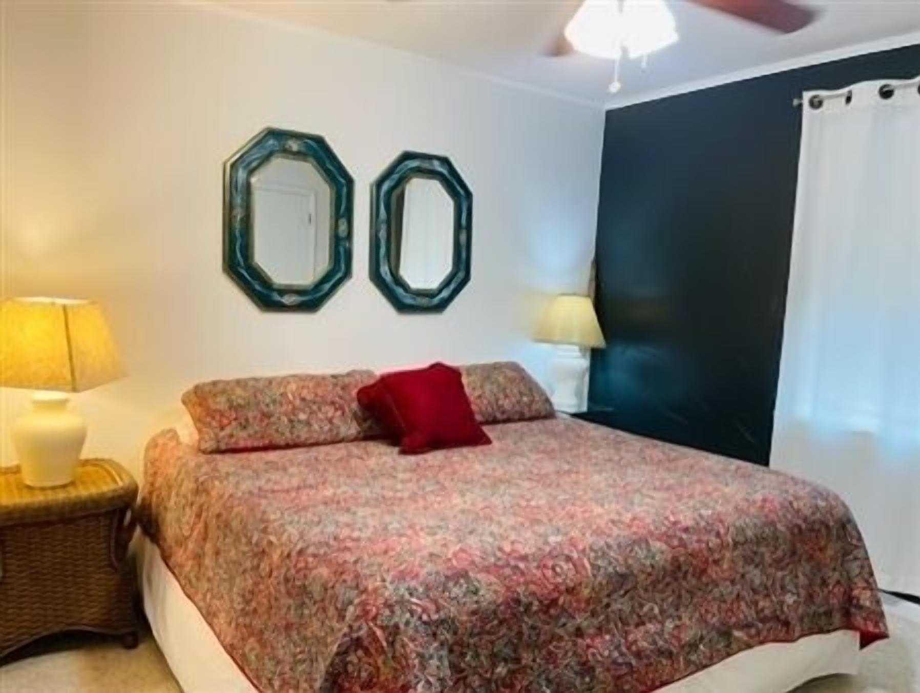 A bedroom with a bed, two wall mirrors, a red pillow, a lamp, and a ceiling fan.